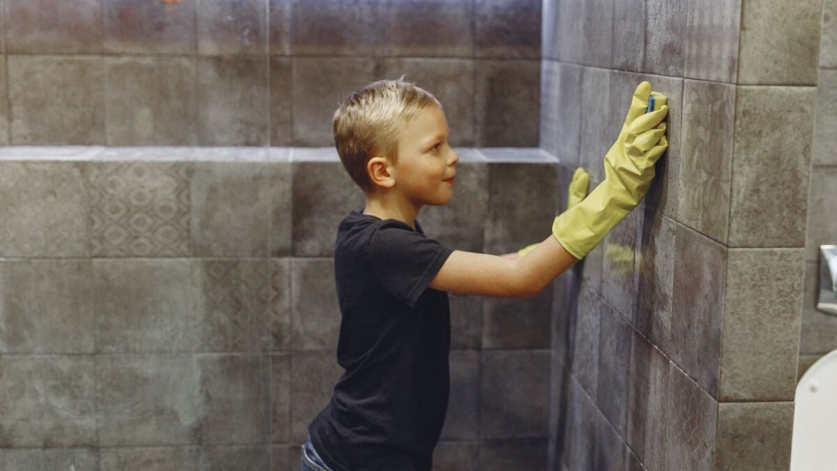 Side view of kid with blond hair in yellow rubber gloves cleaning tile wall in bathroom, illustrating best tile and grout cleaner for hard water