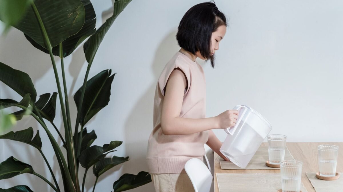 A young girl pours water into glasses at a minimalist dining table with a green indoor plant ne, illustrating best water filter pitcher for hard water