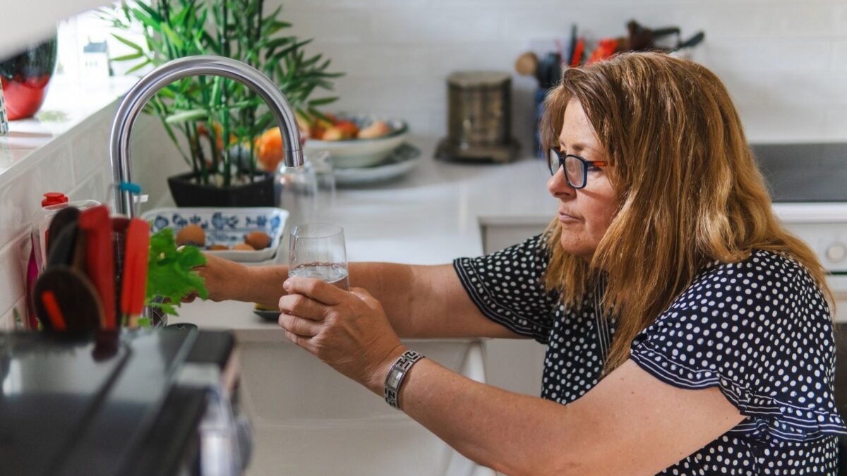 A middle-aged woman filling a glass with water in a modern kitchen setting, illustrating best whole house filter for pfas