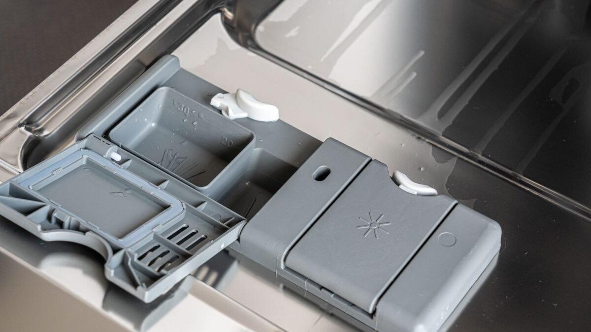 A detailed view of a dishwasher detergent compartment with soap residue, illustrating finish vs cascade hard water